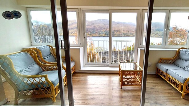 Ein Zimmer mit Korbmöbeln, einem Glastisch und großen Fenstern mit Blick auf einen See und Hügel.
