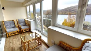 Ein Balkon mit Korbmöbeln, einer Glastür und Blick auf einen See und Hügel.