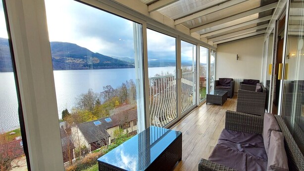 A balcony with a view of a lake and mountains, furnished with wicker sofas and a glass table.