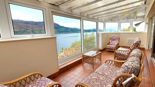 A sunlit balcony with wicker furniture, large windows offering scenic views of hills and water. - Loch Rannoch Highland Club, Highland Lodge 36 (Kinloch Rannoch)