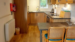 A compact kitchen with wooden cabinets, a built-in stove, a sink, and a dining area with two wicker chairs. - Loch Rannoch Highland Club, Lochside Lodge 7 (Kinloch Rannoch)