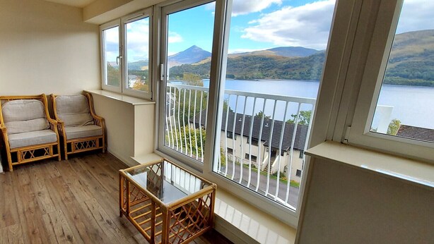 A balcony with two wicker chairs, a glass table, and a view of a lake and mountains. - Loch Rannoch Highland Club, Otter Lodge 1 (Kinloch Rannoch)