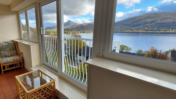 A balcony with two wicker chairs, a small table, and a view of a lake and mountains. - Loch Rannoch Highland Club, Otter Lodge 3 (Kinloch Rannoch)