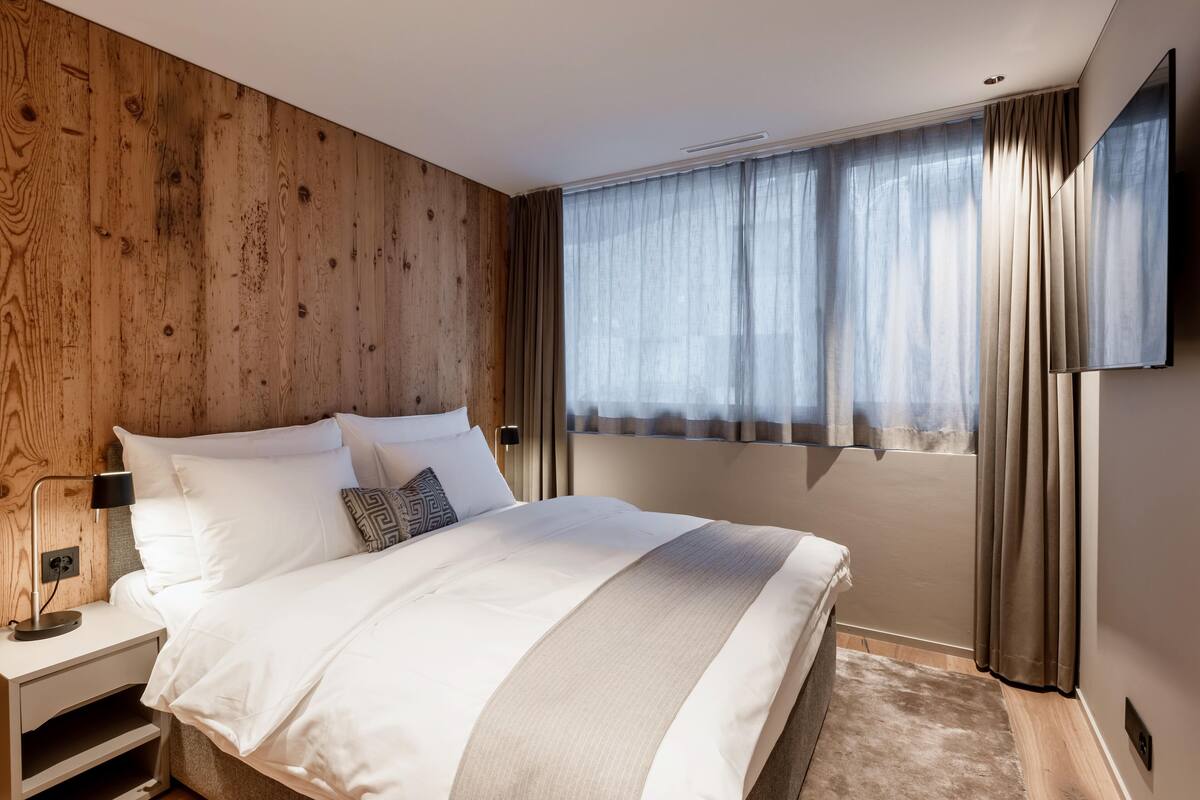 A modern bedroom with a wooden wall, a large bed, a TV, and a window with curtains.