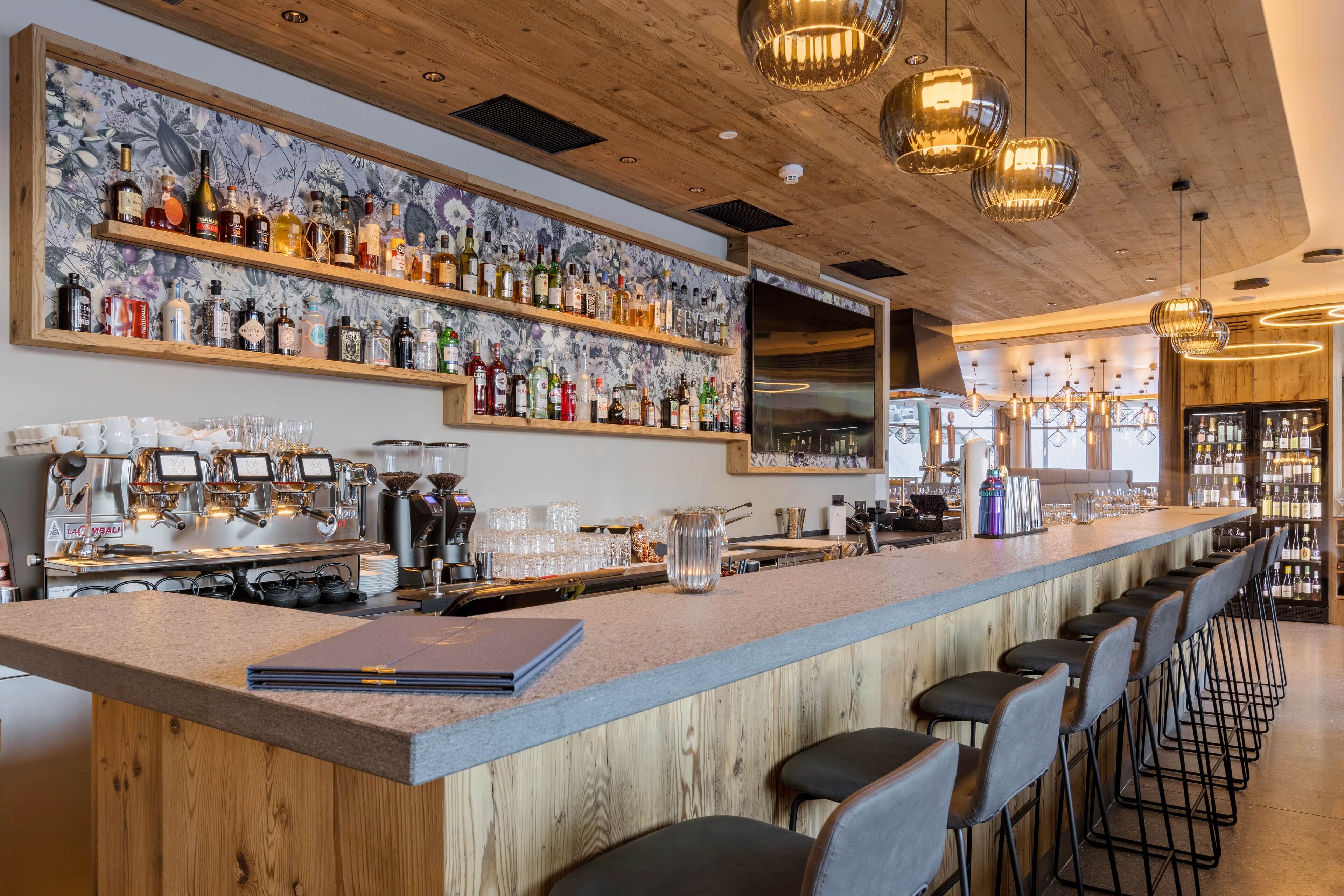 Zona de bar con estantes de madera que exhiben varias botellas, una máquina de café y una larga barra con taburetes.