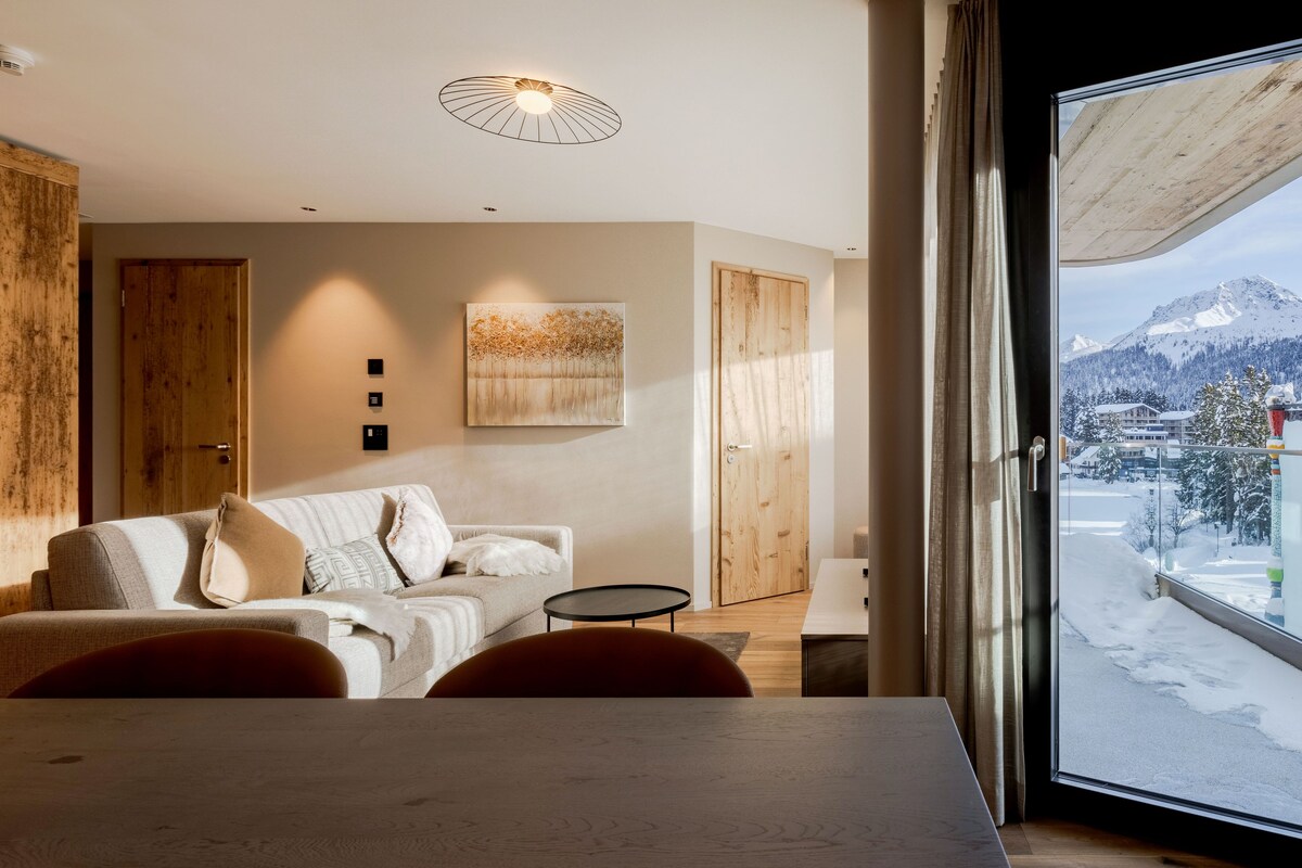A modern hotel room with a large window, a wooden door, a sofa, a small table, and a painting on the wall.