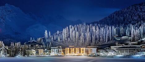A snow-covered mountain town at night with illuminated buildings and a frozen lake.
