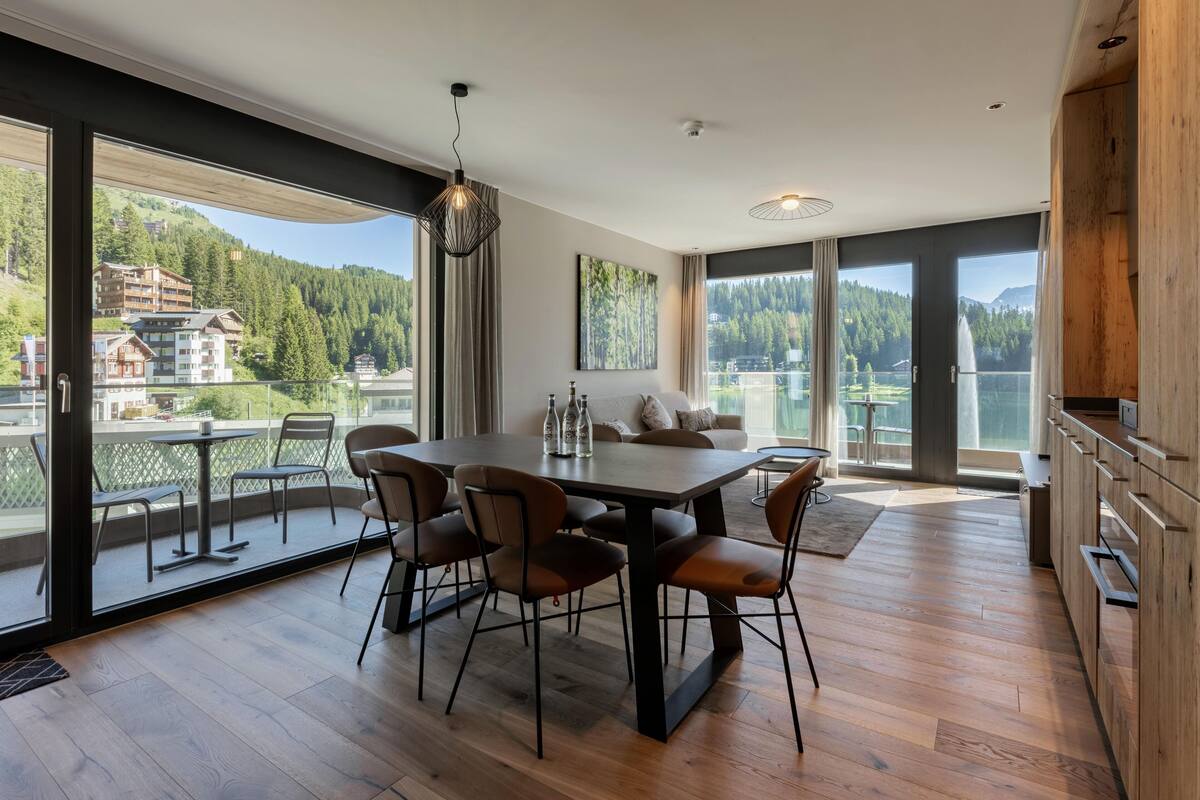 A modern dining area with a large table and chairs, large windows with a view of a mountain and buildings, and a wooden cabinet.