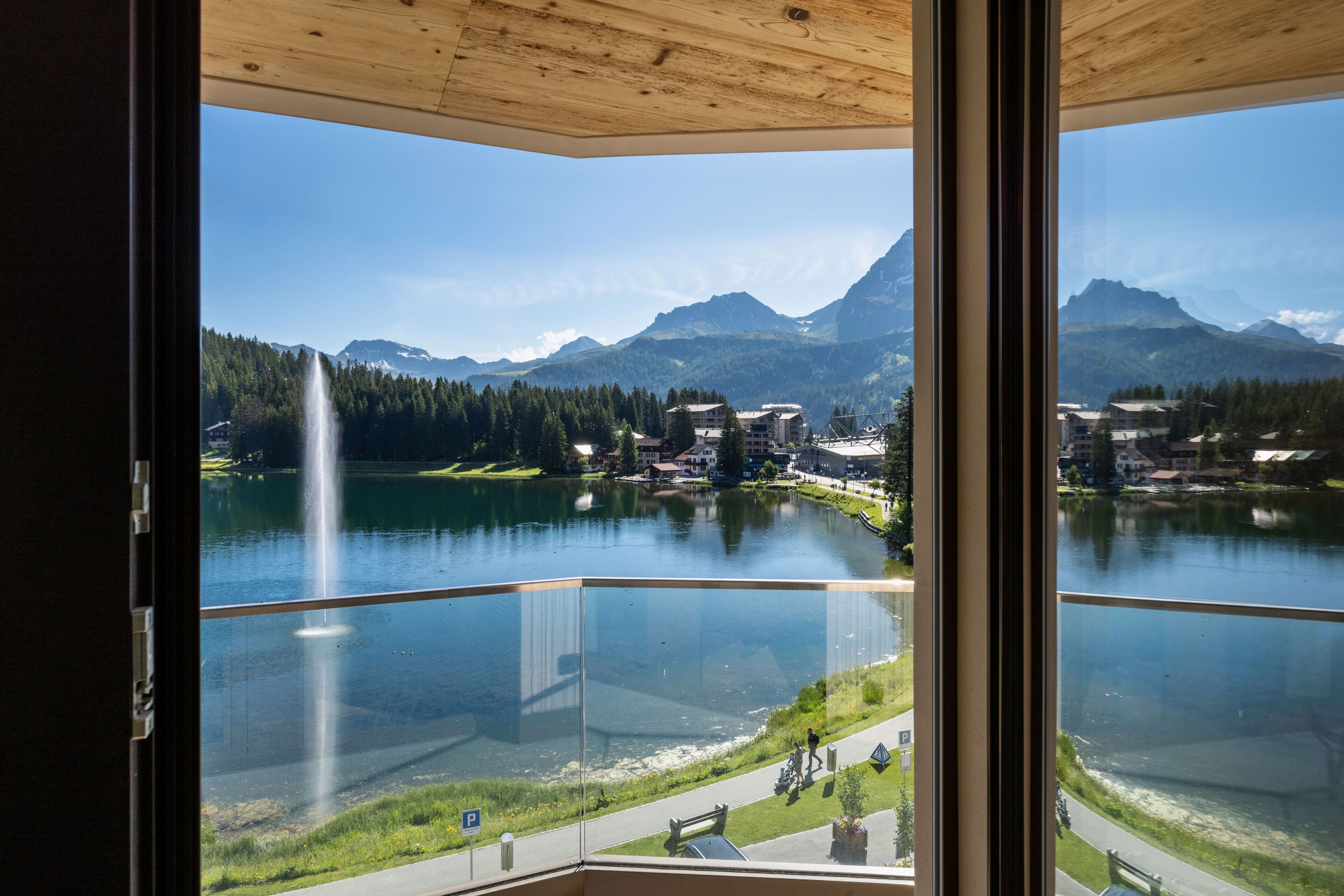 A view from a balcony overlooking a lake with a waterfall, mountains, and buildings in the background.