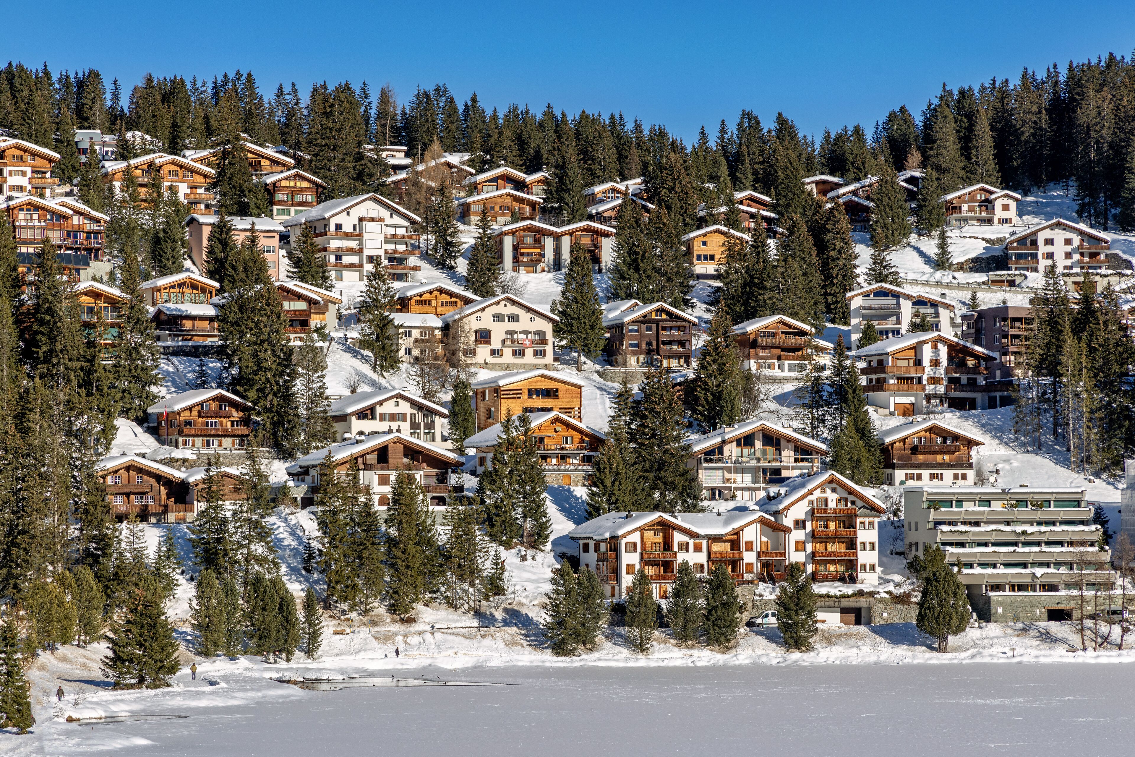 A cluster of log cabins with multiple stories, surrounded by snow-covered ground and pine trees, indicating a winter resort setting.