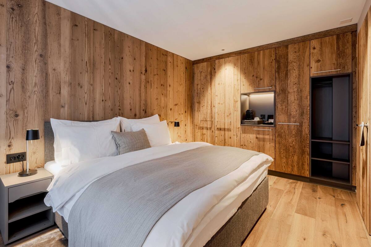 A modern bedroom with a wooden wall panel, a large bed with white bedding, a bedside table with a lamp, and a built-in wardrobe.