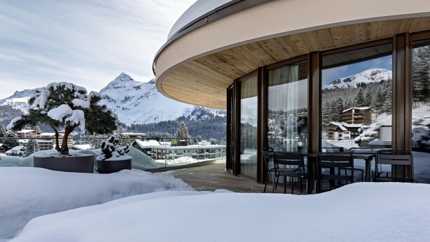 A modern building with large windows, a wooden deck, and a snowy mountain backdrop. - Postresidenz am See (Arosa)
