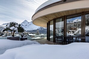 Un bâtiment moderne doté de grandes fenêtres, d’une terrasse en bois et surplombant un paysage montagneux enneigé.