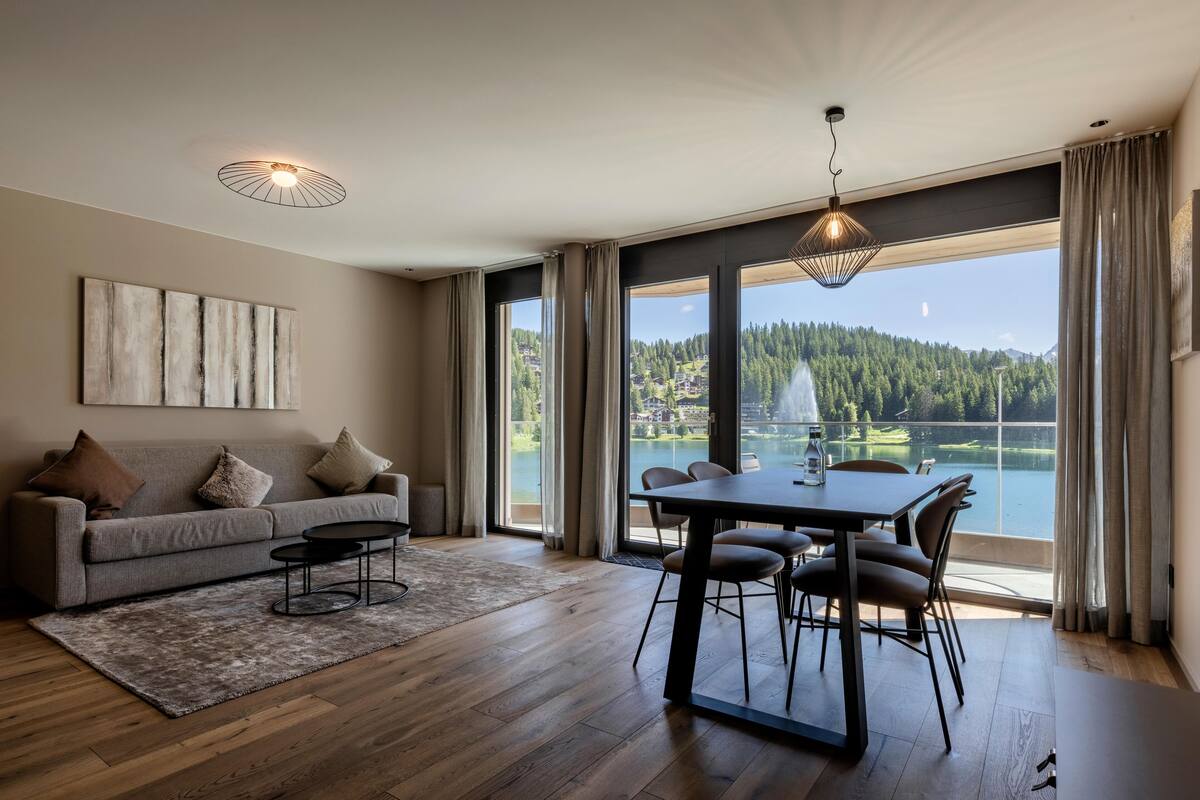 A modern living room with a sofa, coffee table, and dining table. Large windows offer a view of a lake and forest.