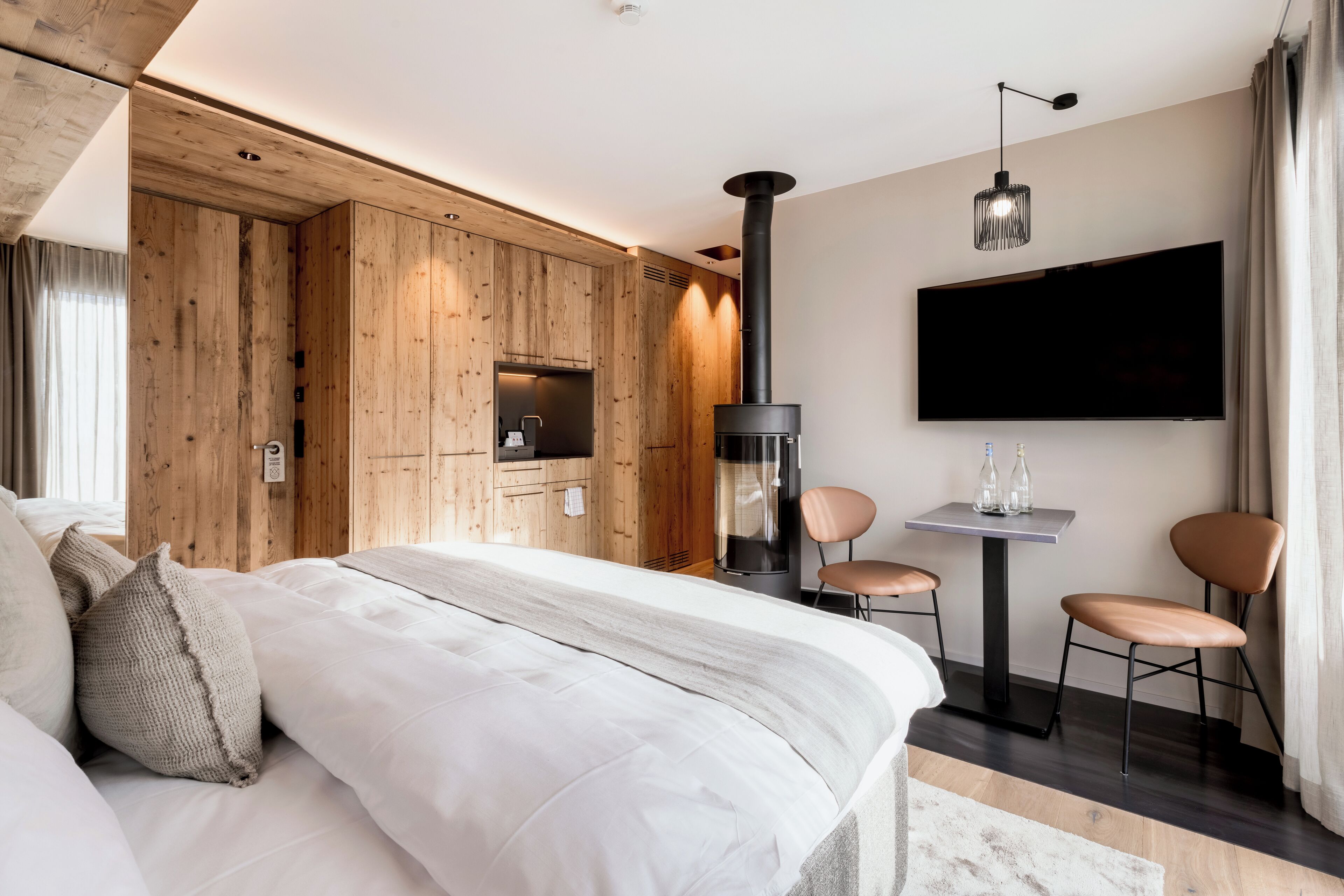 A modern hotel room with a large bed, wooden panel walls, a fireplace, a flat-screen TV, and two chairs.
