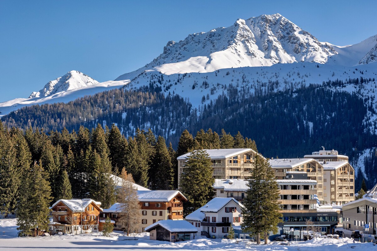 A mountain resort with wooden buildings, a ski lift, and snow-covered peaks.