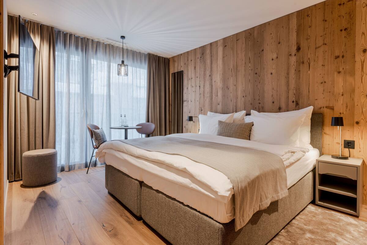 A modern bedroom with a large bed, wooden panel walls, a round ottoman, and a small table with chairs.