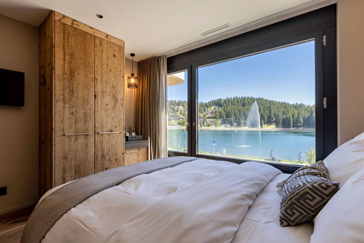 A bedroom with a large window, a wooden wardrobe, and a bed with white bedding.