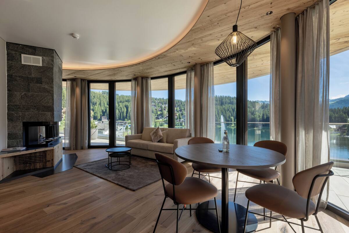 A modern living room with a stone fireplace, a round dining table with chairs, and large windows offering a view of a forested area.