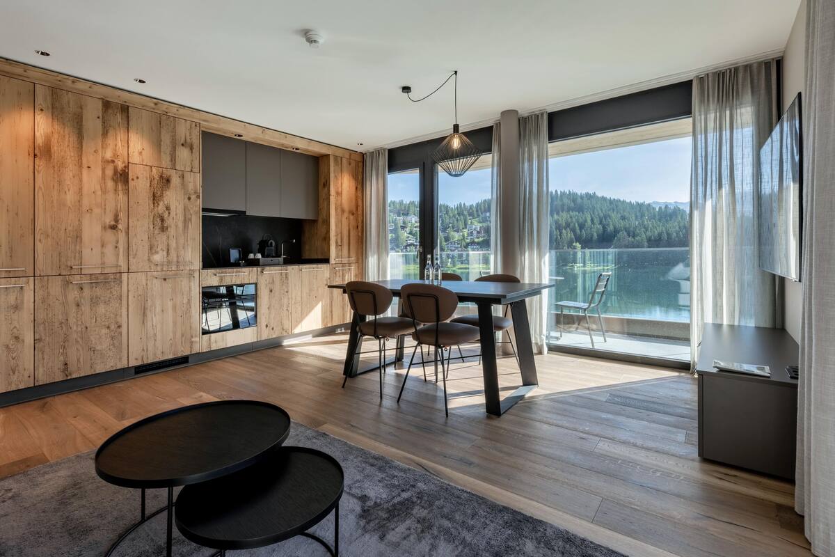 A modern kitchen with wooden accents, a dining area with a table and chairs, and a large window offering a lake view.
