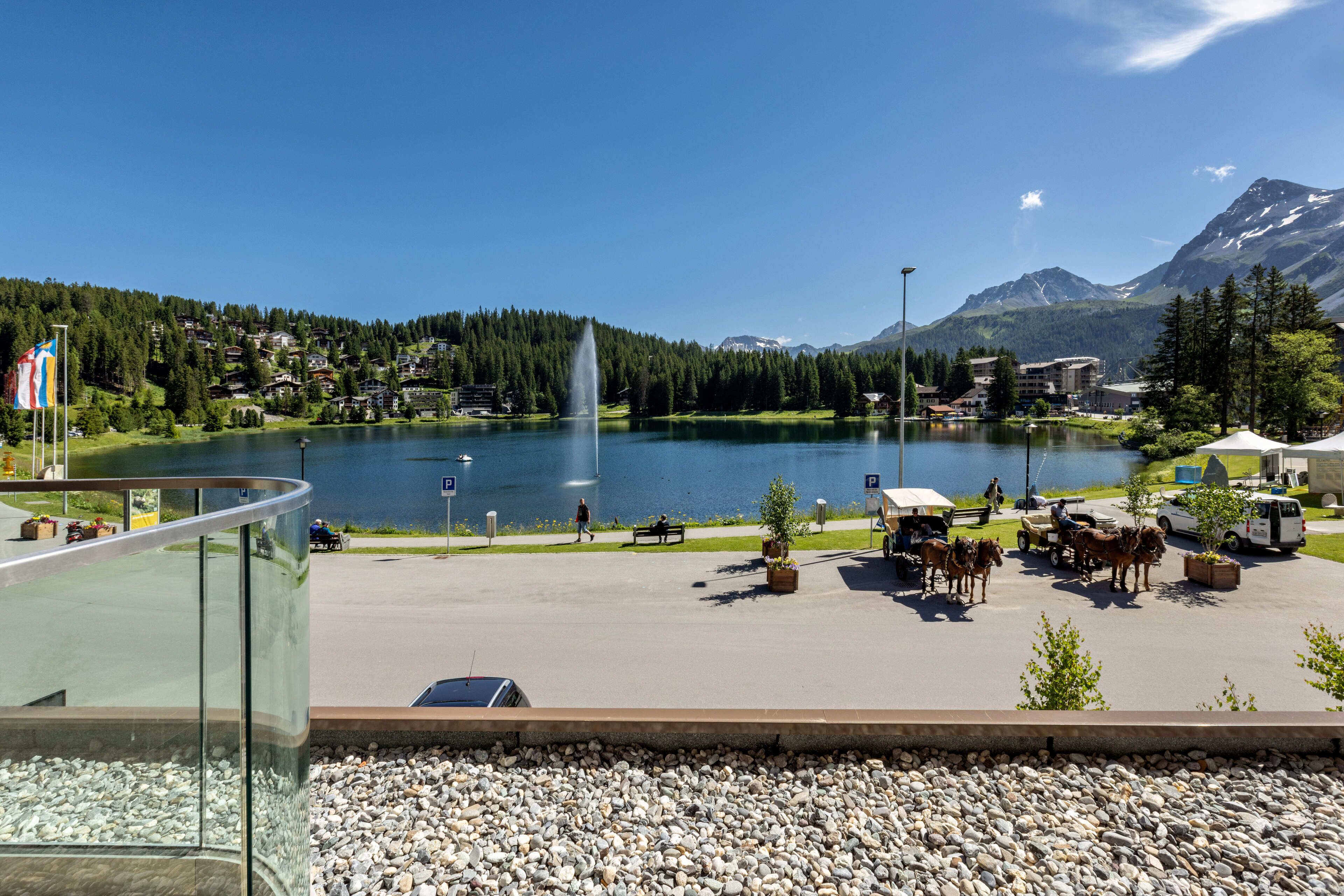 A lakeside view with a fountain, a parking area, and a mountain backdrop.