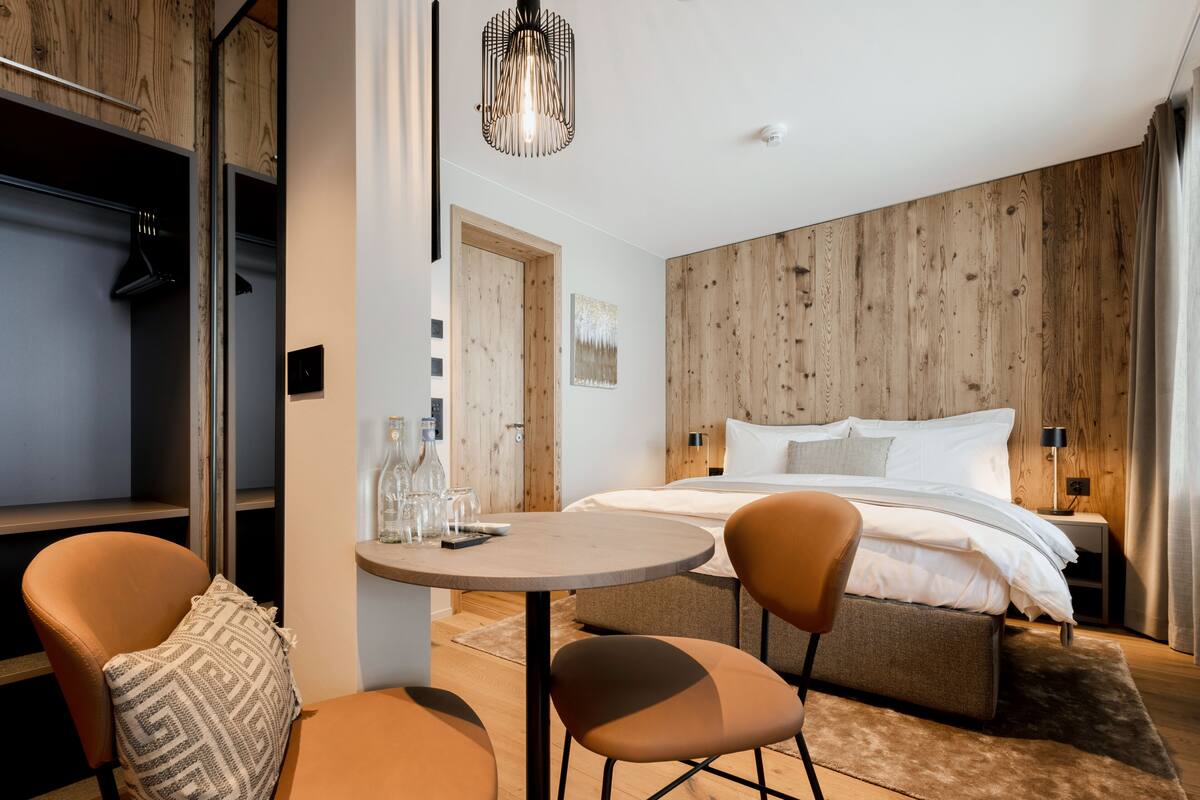 A modern bedroom with a wooden wall, a bed with white bedding, a round table with chairs, and a hanging light fixture.