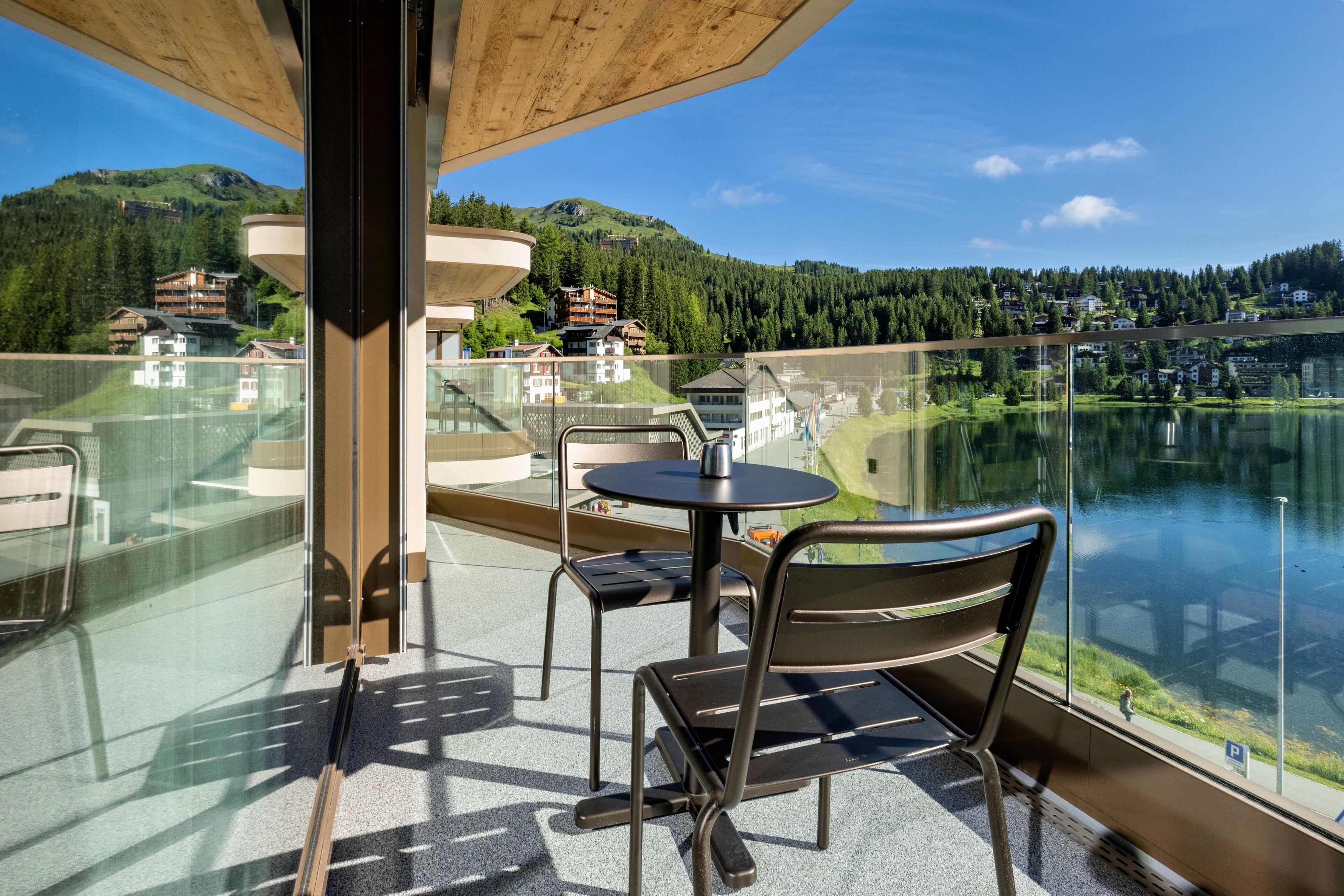 A balcony with a glass railing, a table, and two chairs, overlooking a lake and buildings in a mountainous area.
