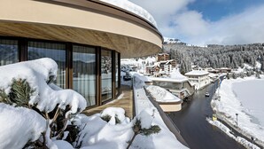 A modern building with large windows and a curved roof, overlooking a snow-covered landscape and a road with vehicles. - Postresidenz am See (Arosa)