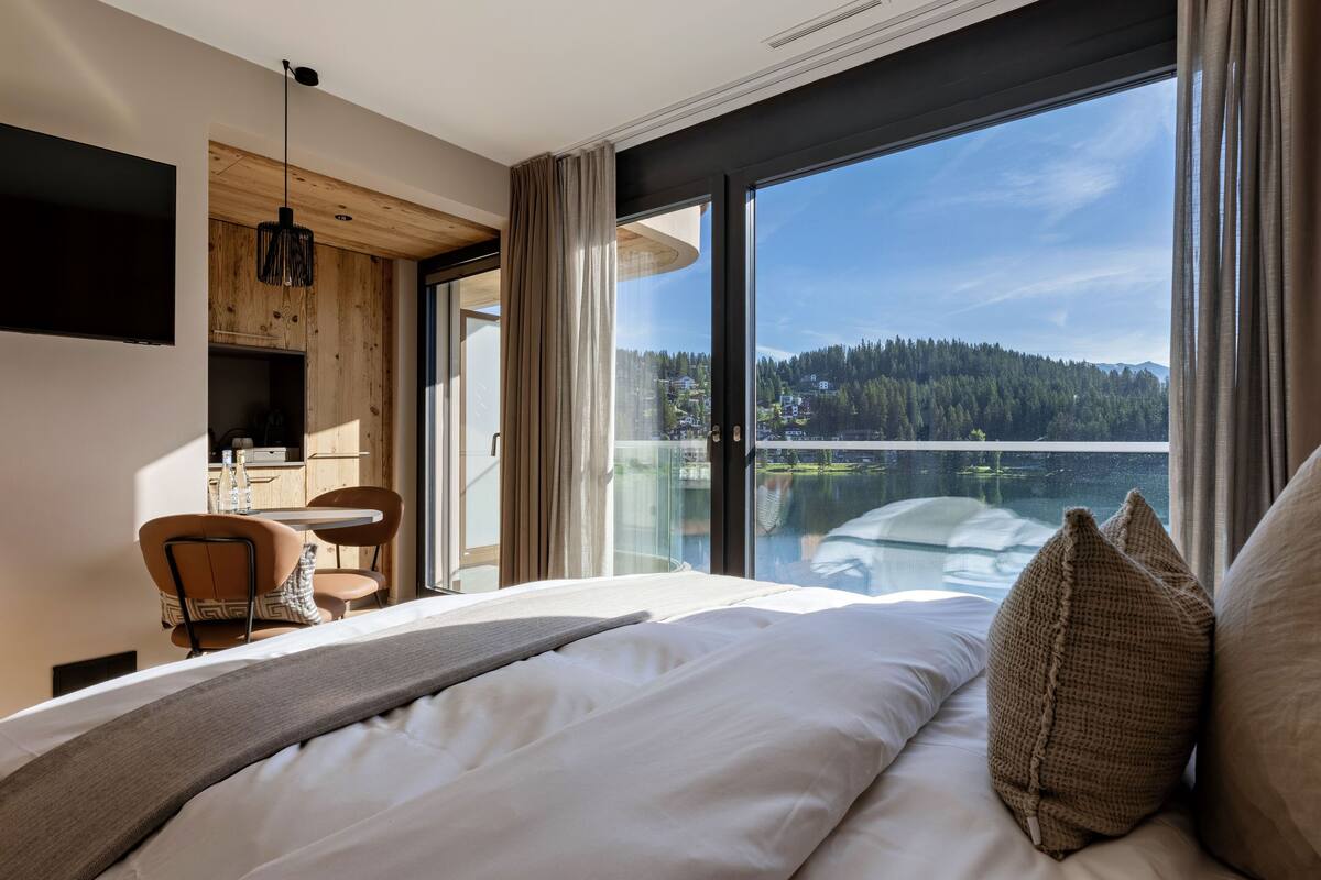 A modern hotel room with a large bed, a dining area, and a view of a lake and forest.