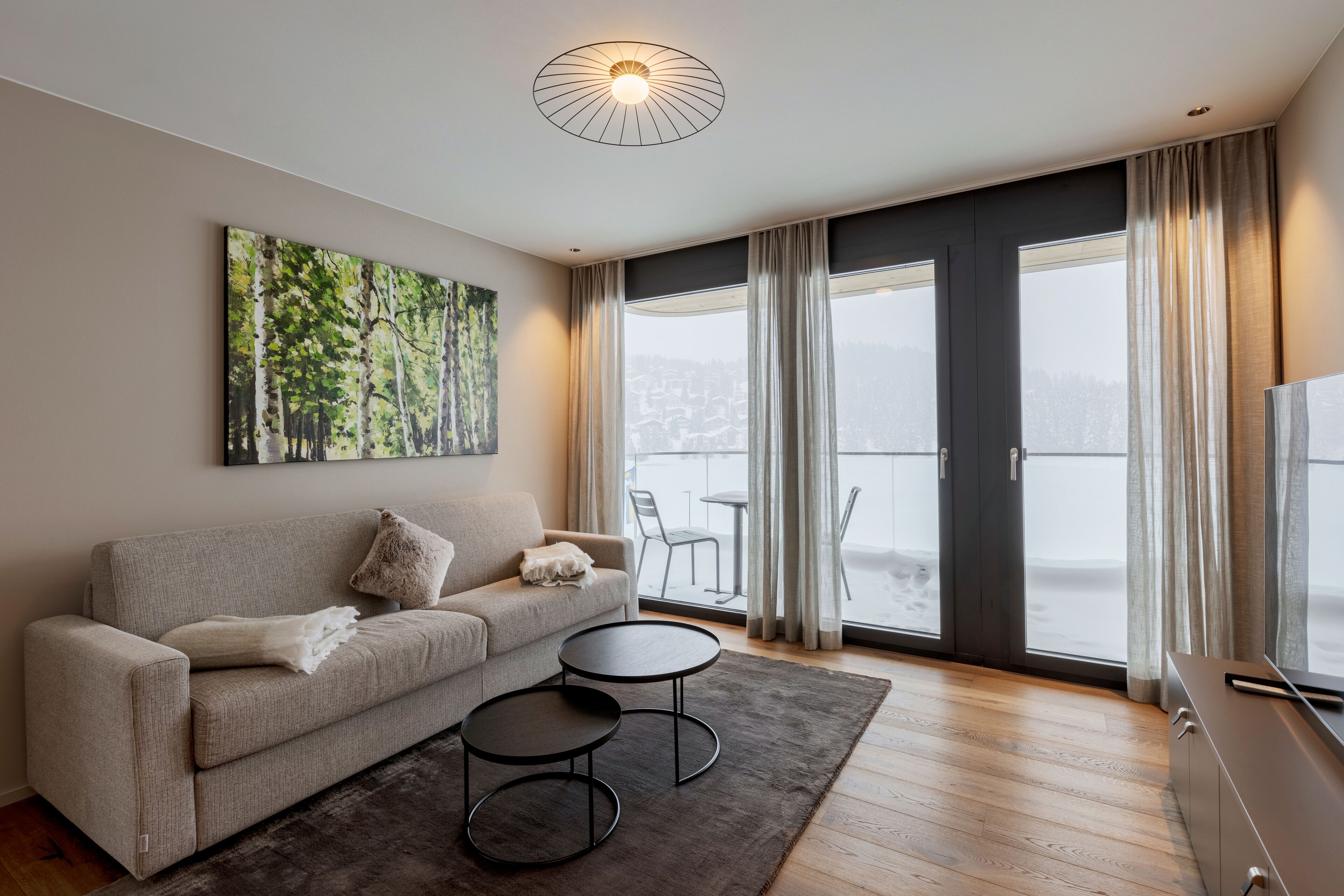A modern living room with a sofa, two round tables, a painting, and a view of a snowy landscape through large windows.