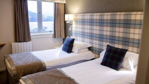 A hotel room with two beds, plaid headboard, a window with curtains, a radiator, and a lamp.