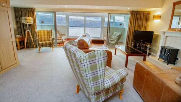 A spacious living room with a large sofa, armchairs, a coffee table, and a television. The room has large windows with curtains and a view of the outdoors.