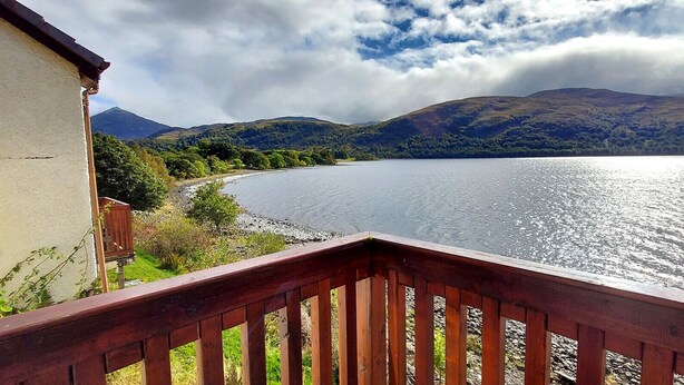 A lakeside view from a balcony with a wooden railing, overlooking a calm lake, surrounded by hills and a partly cloudy sky.