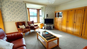 A living room with a sofa, two armchairs, a coffee table, a TV, wooden cabinets, and a window with curtains.