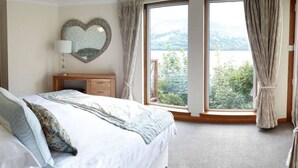Een slaapkamer met een groot bed, een dressoir, een spiegel in de vorm van een hart en uitzicht op een bergachtig landschap door een schuifdeur van glas.