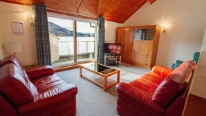Een kamer met twee leren banken, een houten salontafel, een televisie en een glazen schuifdeur die leidt naar een balkon met uitzicht op bergen.