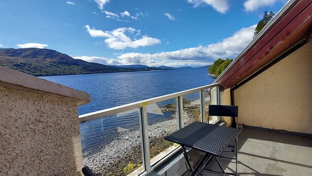 Een balkon met een metalen tafel en stoel, met uitzicht op een meer met bergen in de verte.