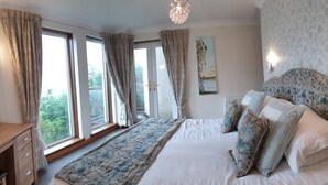 Een slaapkamer met een groot bed, een houten dressoir en uitzicht op groen door de ramen.