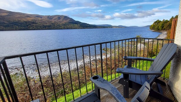 A balcony with a view of a lake and hills, featuring two Adirondack chairs and a small table. - Loch Rannoch Highland Club, Lochside Lodge 6 (Kinloch Rannoch)