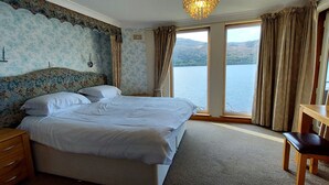 A bedroom with a large bed, a wooden nightstand, a desk, and a view of a lake. - Loch Rannoch Highland Club, Lochside Lodge 6 (Kinloch Rannoch)