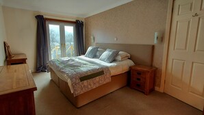 Ein Schlafzimmer mit einem Bett, Nachttischen, einem hölzernen Beistelltisch, einem Fenster mit Vorhängen und einer Tür.