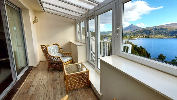 Un balcon offrant une vue sur un lac et des montagnes, aménagé avec des fauteuils en osier et une table en verre.