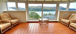 A room with wicker furniture, a glass table, and large windows overlooking a lake and hills.
