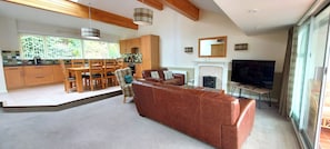 A modern living room with a brown leather sofa, wooden dining table, and chairs. A fireplace is visible, and a television is mounted on the wall.