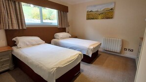 A bedroom with two beds, a window with curtains, a radiator, and a painting on the wall. - Loch Rannoch Highland Club, Pine Martin Lodge 4 (Kinloch Rannoch)