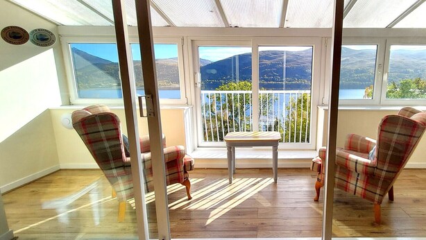 A sunlit room with a view of a lake and mountains, furnished with plaid chairs and a wooden table. - Loch Rannoch Highland Club, Pine Martin Lodge 4 (Kinloch Rannoch)