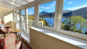 A balcony with a view of a lake and mountains, furnished with plaid chairs and a small table. - Loch Rannoch Highland Club, Pine Martin Lodge 4 (Kinloch Rannoch)