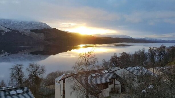 A lakeside view with a sunset, snow-capped mountains, and a row of buildings with sloped roofs.