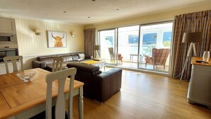 A spacious living area with a dining table, sofa, and a view of the outdoors through large sliding glass doors. - Loch Rannoch Highland Club, Pine Martin Lodge 4 (Kinloch Rannoch)