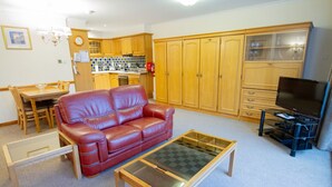A living room with a red leather sofa, a glass-top coffee table, wooden cabinets, a television, and a kitchen area in the background. - Loch Rannoch Highland Club, Red Squirrel 4 (Kinloch Rannoch)